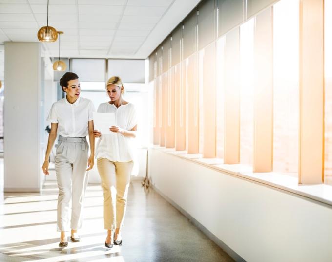 Two businesswoman walk down an office building’s sunlit hallway.