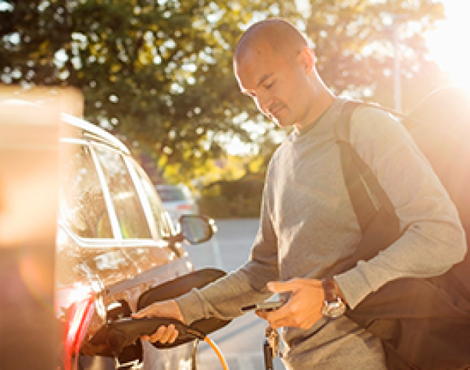 Middle aged male plugging a charger connector into his electric vehicle while looking at his phone on a sunny day.   