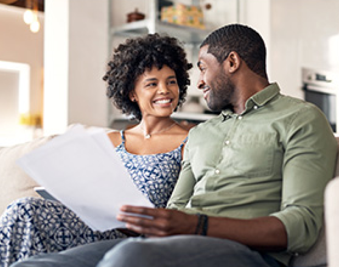 A woman wearing a blue and white dress and man wearing an olive button-up with jeans are sitting down, looking into each other’s eyes and smiling. The man is holding up papers.