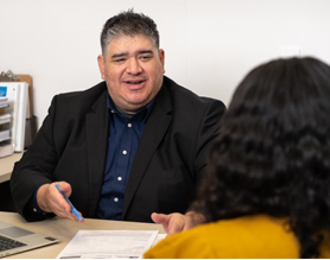 Smiling customer service representative is sitting down holding a pen in hand with paperwork on the table assisting a customer.