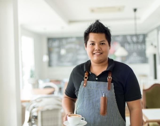 Individual wearing an apron standing with a cup of coffee inside a cafe.