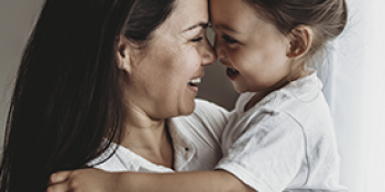 A woman with long, dark brown hair holding up and embracing a young girl with brown hair in a bun. Both are wearing white collared shirts and smiling with their foreheads touching.