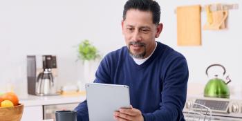 Man seated in kitchen looking at a computer tablet.